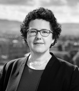 Black and white headshot of Joanna Baker. She looks directly at the camera, smiling, wearing glasses and a blazer.