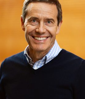 Peter Maniura smiling at the camera against a bright orange background.