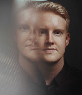 A man looks towards the camera against a black background. The left side of the image has been distorted and a faint copy of the man's face is visible.