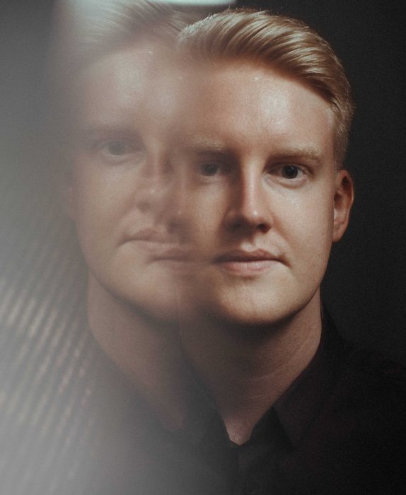 A man looks towards the camera against a black background. The left side of the image has been distorted and a faint copy of the man's face is visible.