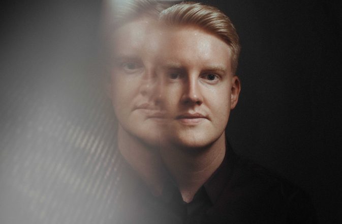 A man looks towards the camera against a black background. The left side of the image has been distorted and a faint copy of the man's face is visible.