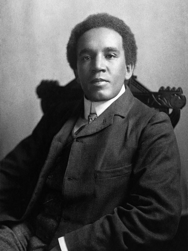 Samuel Coleridge Taylor sits on a chair. The photo is in black and white and he is in formal ware.