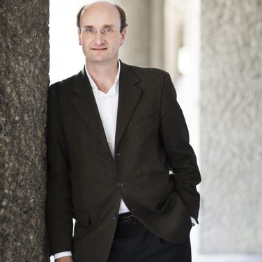 Conductor Andrew Manze stands in a suit, facing the camera, leaning against a wall to his right.