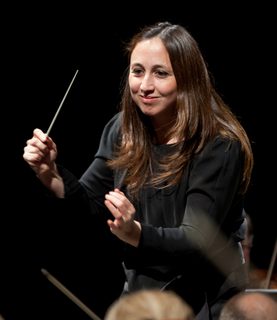 Conductor Joana Carneiro smiles as she leads the orchestra, her hand raised mid-gesture.
