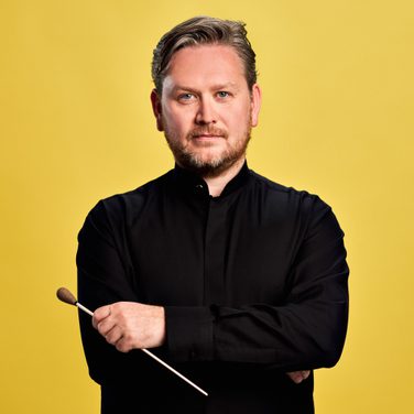 Conductor Jonathan Bloxham stands with his arms crossed, looking into the camera with a light smile, in front of a yellow background.