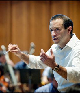 Conductor Mark Wigglesworth, captured from the side mid-conducting, his arms raised with a baton held in his right hand.