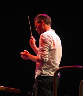 Robert Ames conducts he wears a white shirt and holds a baton facing away from camera.