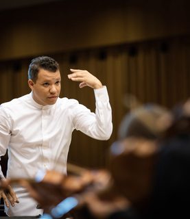 Conductor Ryan Bancroft directs the orchestra, his left arm raised, in a dynamic conducting pose.