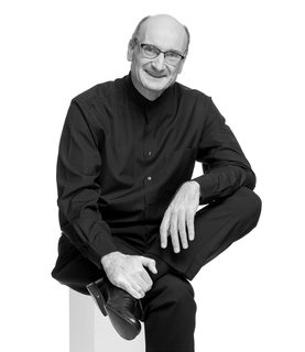 Andrew sits and crossed one leg across the other. He smiles and wears glasses and a black shirt.