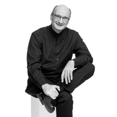 Andrew sits and crossed one leg across the other. He smiles and wears glasses and a black shirt.
