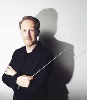 Conductor Simon Crawford-Phillips poses with arms crossed, holding the baton, looking directly into the camera.