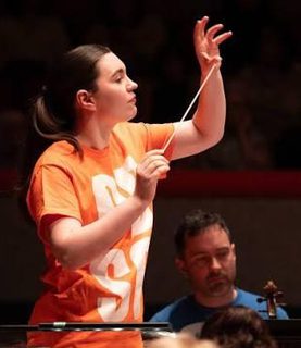 Tess holds a baton and conducts wearing an orange tshirt.
