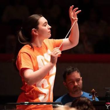 Tess holds a baton and conducts wearing an orange tshirt.