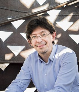 Conductor Václav Luks sits leaning forward, arms resting on his knees, smiling as he looks at the camera.