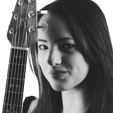 A black and white close up of Devin. She holds her guitar upright and you can see the top of it. She has straight dark hair.