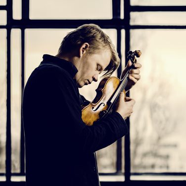 Violinist Pekka Kuusisto, eyes closed, holds his violin close to his face, plucking the strings with his fingers, as he looks down.