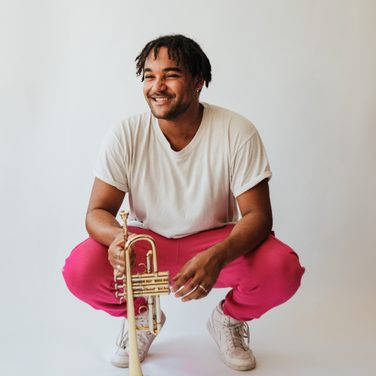 Aaron Azunda Akugbo squats down, holding his trumpet, laughing.