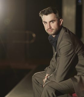 Tenor Anthony Gregory sits onstage, facing the camera against a backdrop of bright stage light.