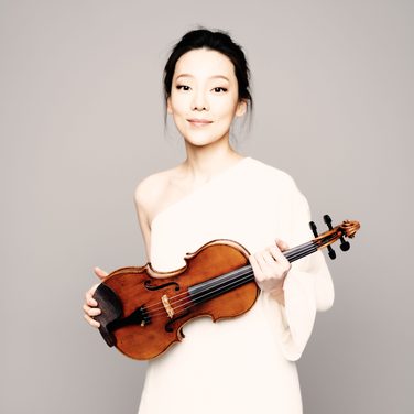 Clara-Jumi Kang, holding her violin, smiles at the camera in a white dress.