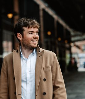 Timothy Ridout, wearing a smart coat and shirt, looks to the right smiling.