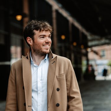 Timothy Ridout, wearing a smart coat and shirt, looks to the right smiling.