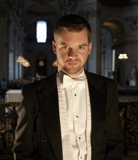 Bass-baritone Edward Grint stands, head slightly tilted down, gazing directly up at the camera.