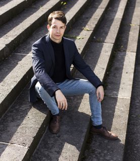 Bass-baritone Florian Störtz sits outdoors on large steps, arms resting on his knees as he looks up at the camera.