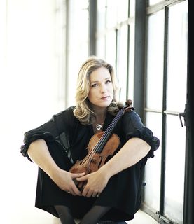 Violinist Isabelle van Keulen sits on the floor with legs angled, arms resting on her knees, holding her violin between her legs.