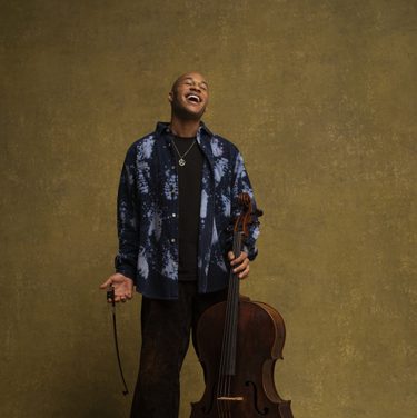 Sheku Kanneh-Mason stands with his cello, tilting his head back in laughter.