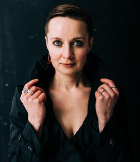 Mezzo-soprano Katie Bray looks directly into the camera, framing her face with both hands holding the collar.