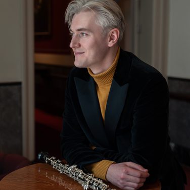 Oboist Ivan Podyomov sits at a small table, his arms crossed and resting on the table, with his oboe lying in front of him, smiling as he looks towards the left of the image.