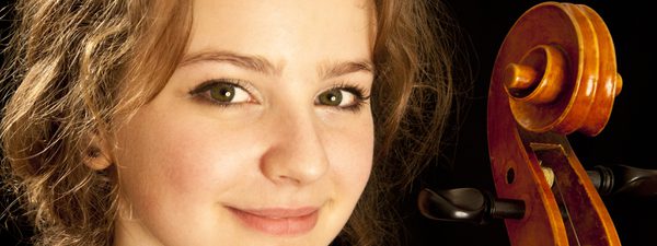 A woman in a dark dress looks towards the camera at a slight angle. She looks with a closed smile and is against a back background. The scroll of a cello is visible.