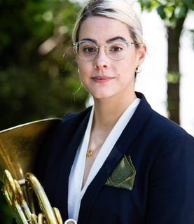 Horn player Lauren Reeve-Rawlings holds her horn in front of her, looking directly at the camera with a gentle smile.