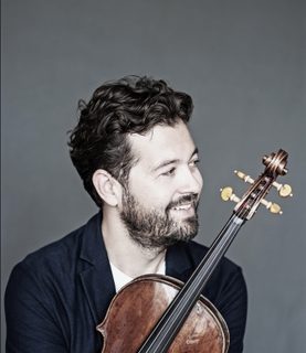 Violinist Lawrence Power sits, smiling and looking to his left, holding his violin close to his chest.