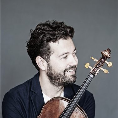 Violinist Lawrence Power sits, smiling and looking to his left, holding his violin close to his chest.