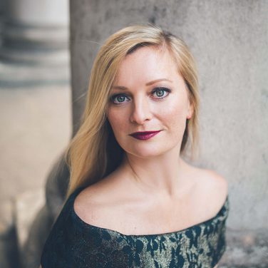 Soprano Lucy Crowe poses sitting down in an elegant dress, looking directly at the camera.