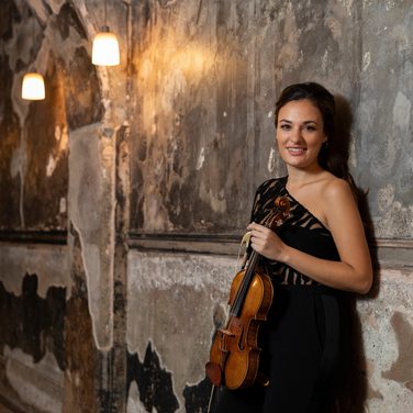 Violinist Nicola Benedetti leans against a wall, holding her violin.