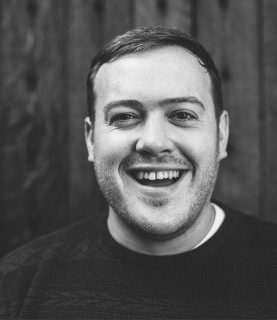 Paul smiles and laugh towards the camera he wears a black jumper.