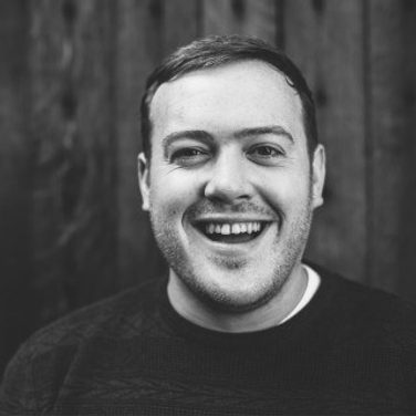 Paul smiles and laugh towards the camera he wears a black jumper.