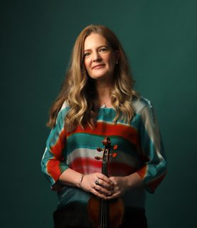 Violinist Rachel Podger stands, smiling at the camera, holding her violin in front of her.
