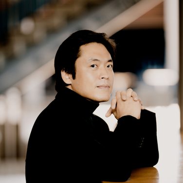 Sunwook Kim rests his arms on a piano, looking at the camera.