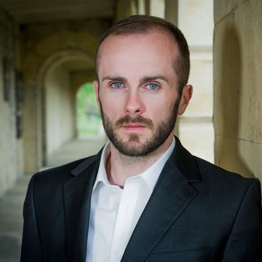 Tenor Thomas Walker stands in the hallway of an old building, leaning against a wall, looking directly into the camera.