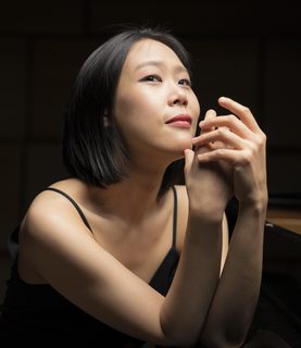 Pianist Yeol Eum Son poses leaning against a piano, her elbows resting on its edge, one hand cradling the other in mid-air, while she tilts her head back and gazes upwards.