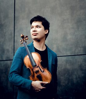 Violinist Aylen Pritchin stands, looking into the distance towards his right, holding his violin in front of him.
