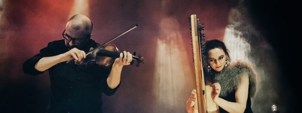 A woman and man are playing harp and violin respectively. They stage against an active background of smoke and coloured stage lighting streaming through.