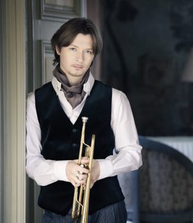 Trumpeter Sergei Nakariakov leans in a doorway, holding his trumpet in front of him.