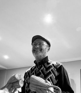A black andwhite photo of Mat who wears glasses and a flat cap holding some percussion instruments