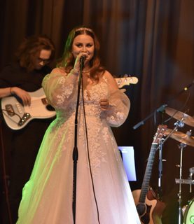 Erin Highton sings into a mic during a performance. She wears a long whitedress.