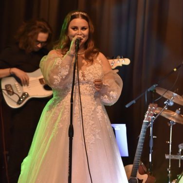 Erin Highton sings into a mic during a performance. She wears a long whitedress.