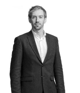 Gregory stands and looks at the camera. He wears a black suit and white shirt.